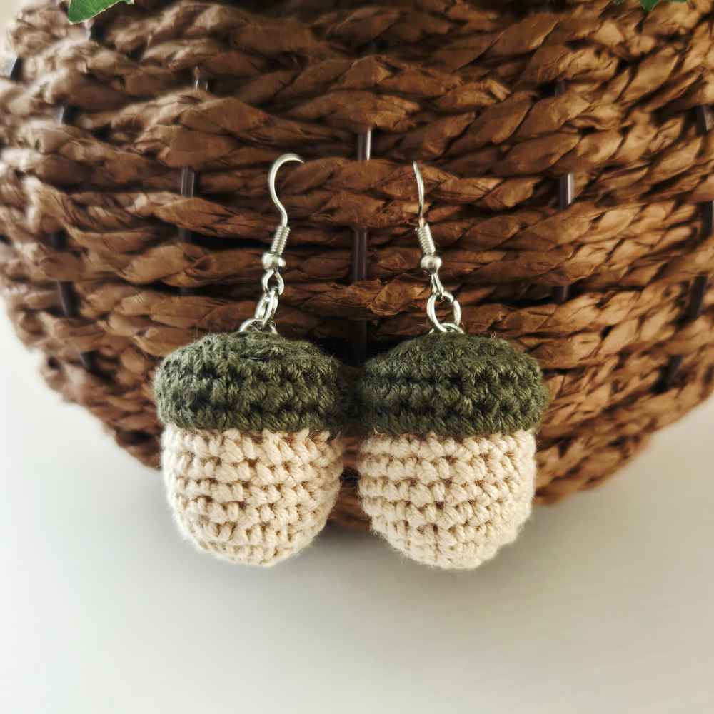 A pair of crocheted acorn dangle earrings with dark olive green tops and cream-colored bodies, hanging from silver fishhook wires in front of a brown woven basket.