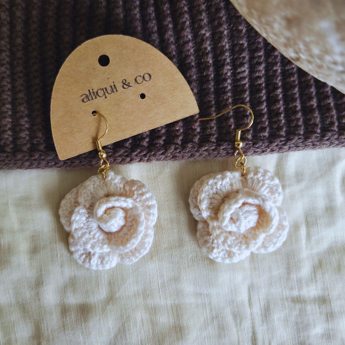 White flower crochet earrings on a brown textured surface with 'aliqui & co' label.