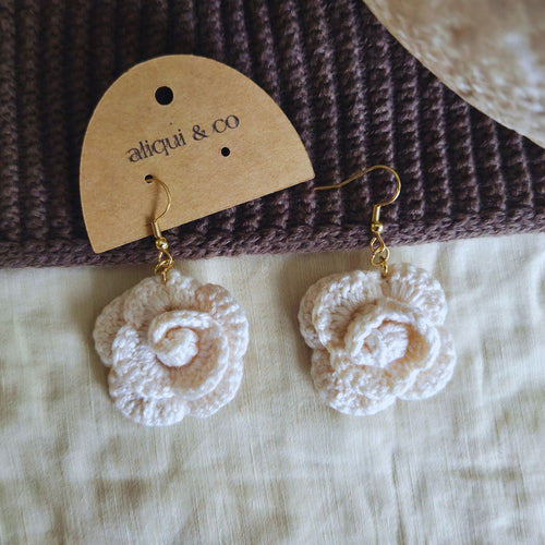 White crochet earrings on a brown textured surface with 'aliqui & co' label.