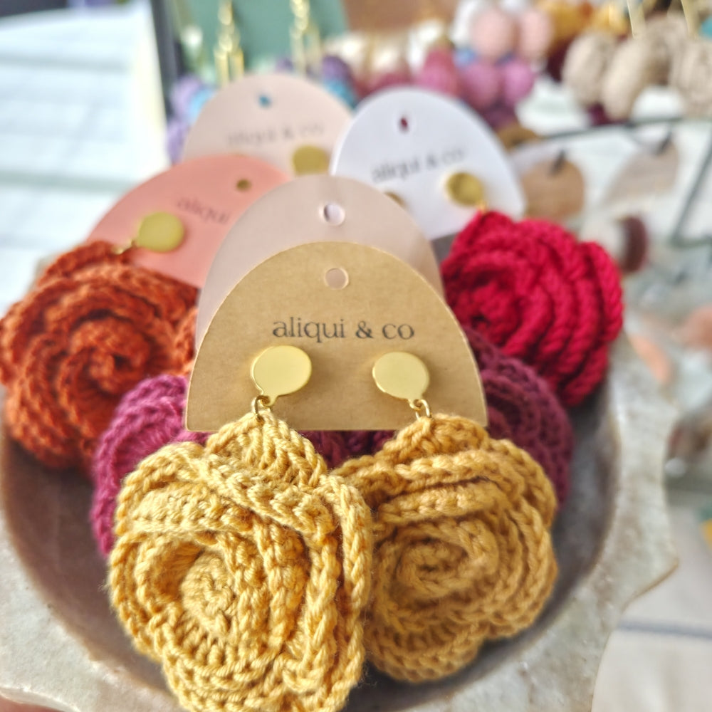 Crocheted flower earrings on a display card with 'aliqui & co' branding, surrounded by other colorful earrings.