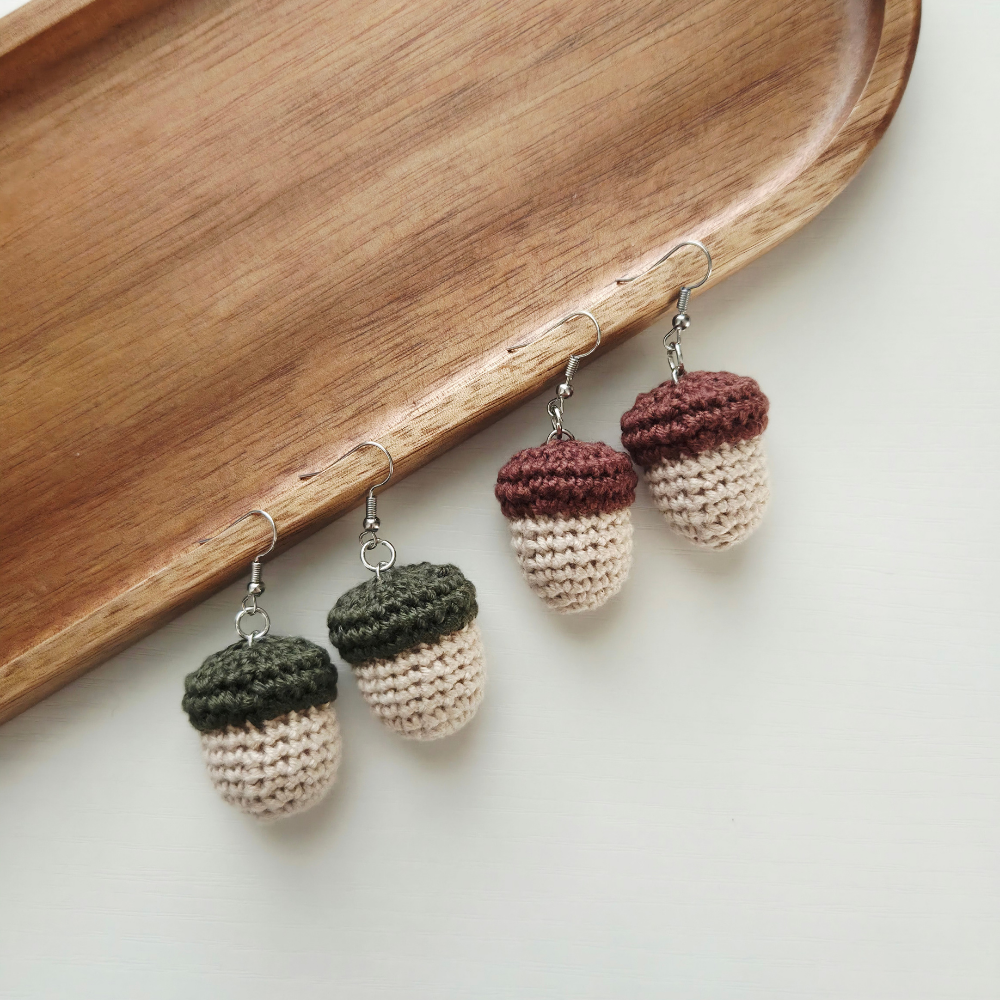 Four crocheted acorn-shaped dangle earrings with silver hooks, displayed on a curved wooden tray against a light off-white background.