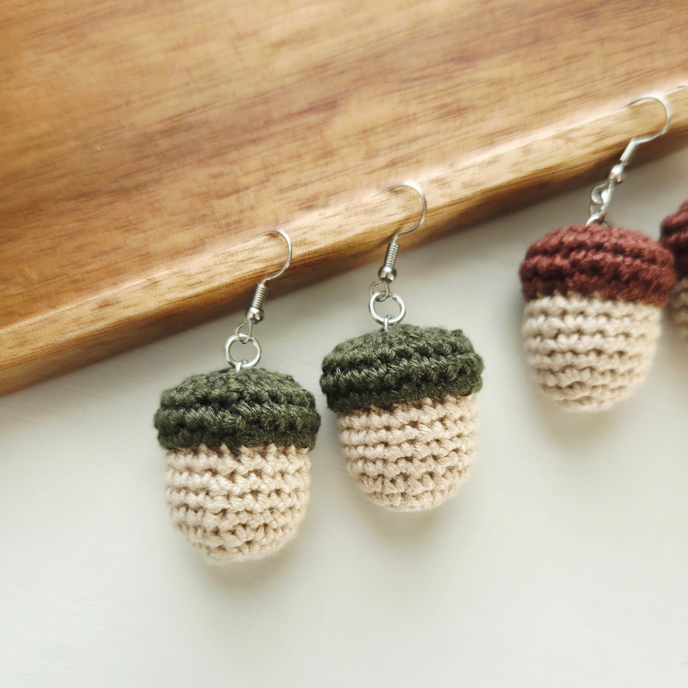 Crocheted acorn dangle earrings hanging from a wooden surface, featuring textured green and beige lower halves with dark brown and beige top halves, attached to silver fishhook ear wires.