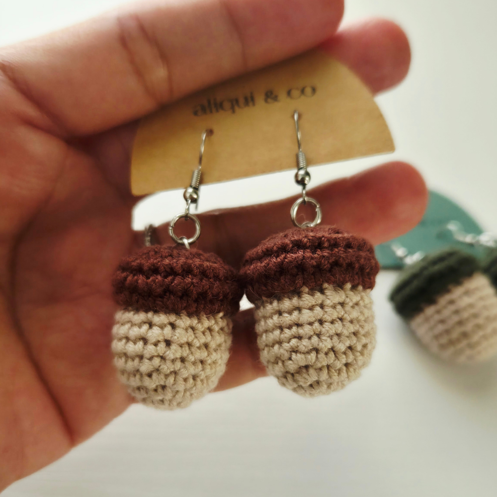 A hand holds a pair of crocheted acorn-shaped dangle earrings with silver fishhook wires. The earrings have dark brown caps and cream-colored bodies, attached to a tan paper display card that reads 'aliqui & co'.