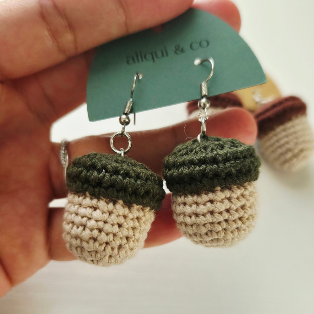 Crocheted acorn drop earrings held in a hand, featuring a dark green top and a cream-colored bottom, with silver fishhook wires. A green paper tag reading 'aliqui & co' is attached to the hooks.