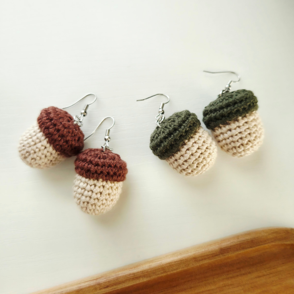 Two pairs of crocheted acorn earrings with silver fishhook wires, displayed on a light cream surface with a wooden edge visible in the bottom right corner. Two pairs are brown and cream, and one pair is dark green and cream.