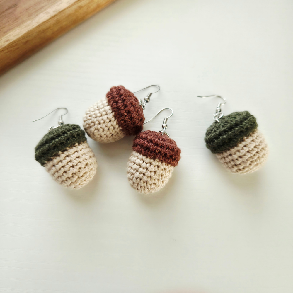 Two handmade crochet acorn dangle earrings with silver fishhook wires, displayed on a light cream surface with a wooden board edge in the top-left corner.