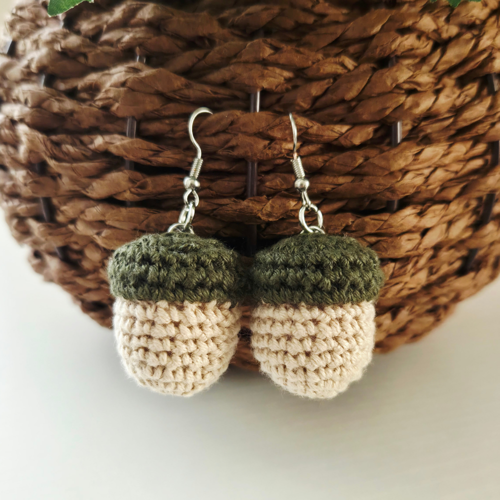 A pair of crocheted acorn dangle earrings with dark olive green tops and cream-colored bodies, hanging from silver fishhook wires in front of a brown woven basket.