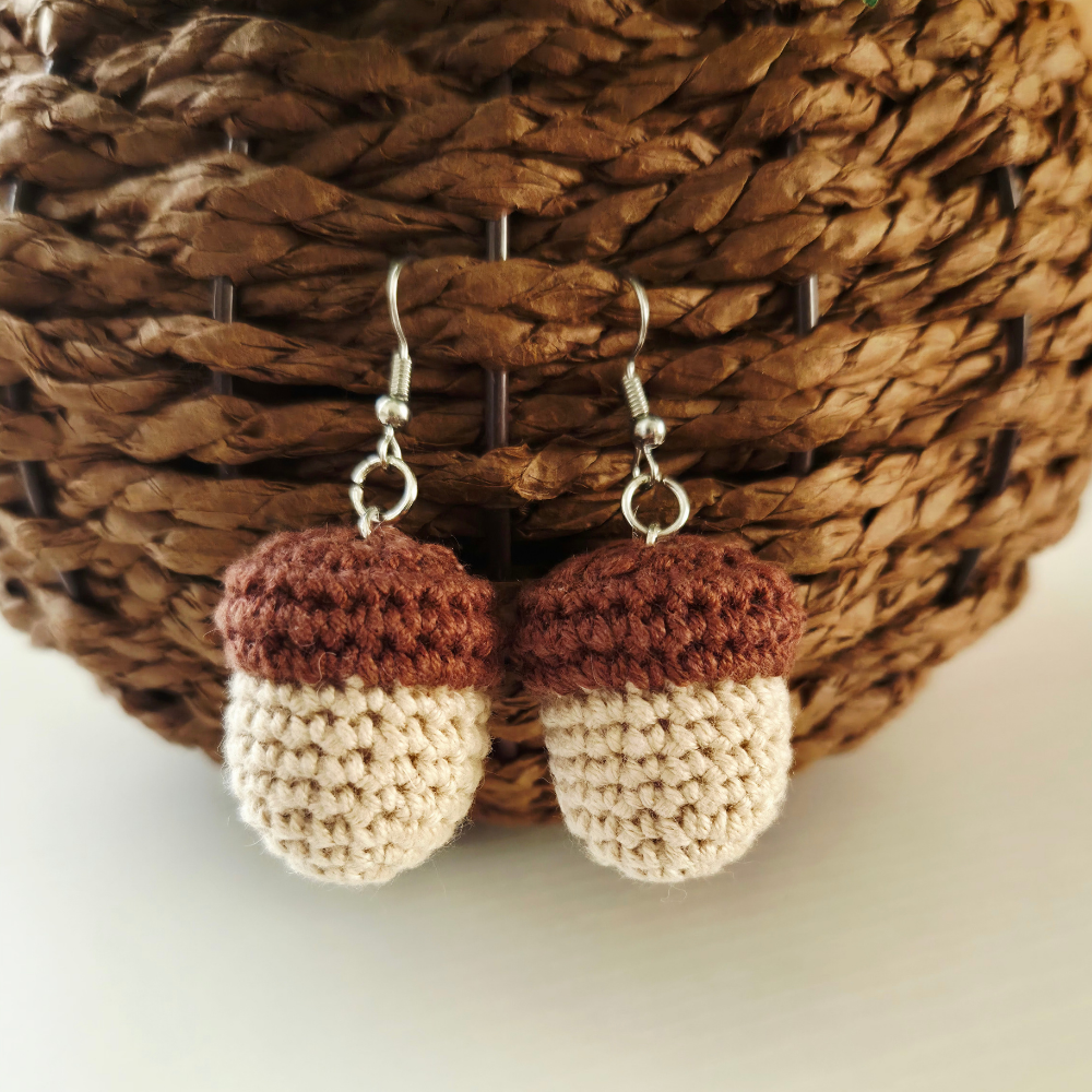 A pair of crocheted acorn-shaped dangle earrings with silver fishhook backings, displayed against a brown woven wicker basket.