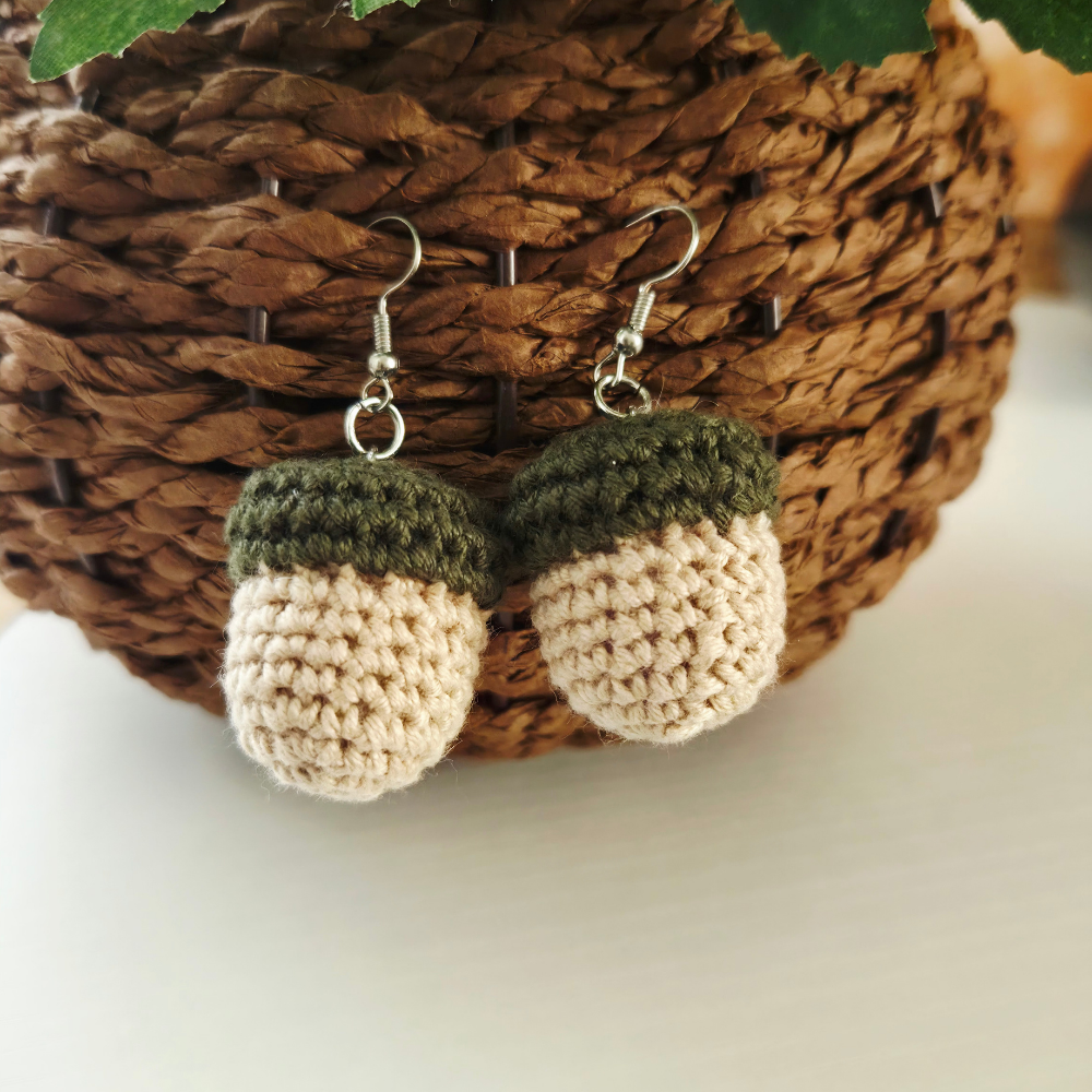 A pair of handmade crochet acorn dangle earrings with silver fishhook wires, resting on a brown woven basket.