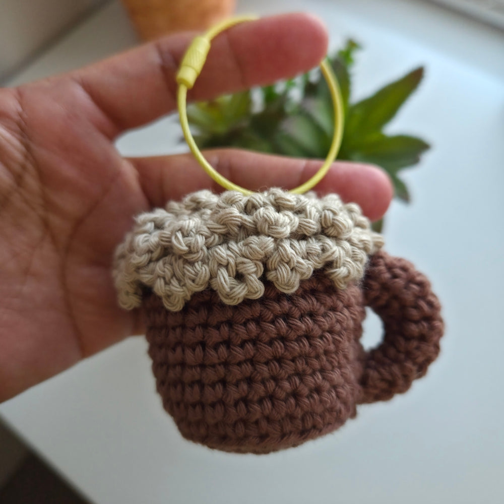 A hand holding a crocheted mug-shaped charm or keychain with a yellow ring, featuring a brown body and a beige textured rim, against a blurred indoor background with a plant.