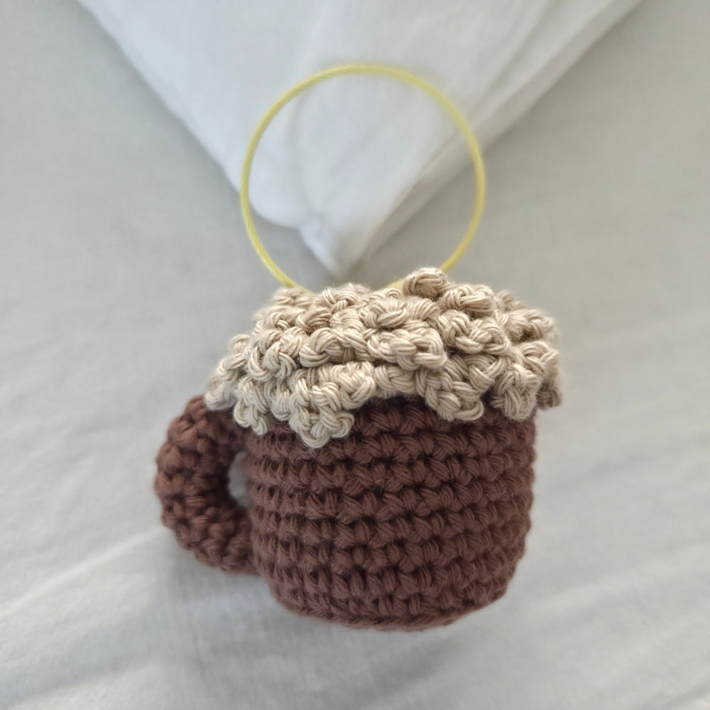 A small crocheted mug-shaped ornament or charm in brown yarn with a beige foam top and a yellow loop at the top, photographed on a soft white fabric background.