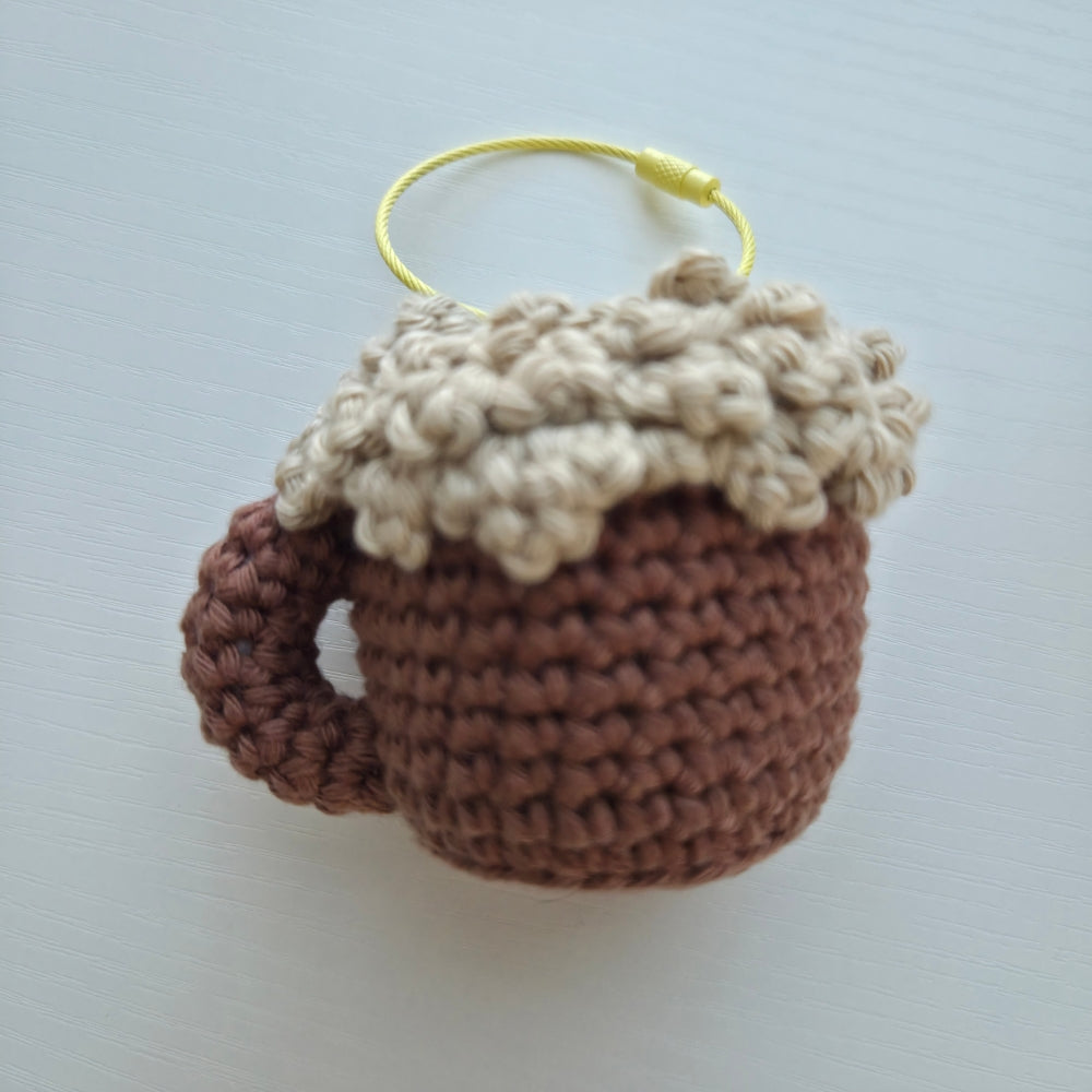 A crocheted beer mug keychain or charm in brown and beige yarn foamy top with a yellow hanging loop, shown on a light gray background.