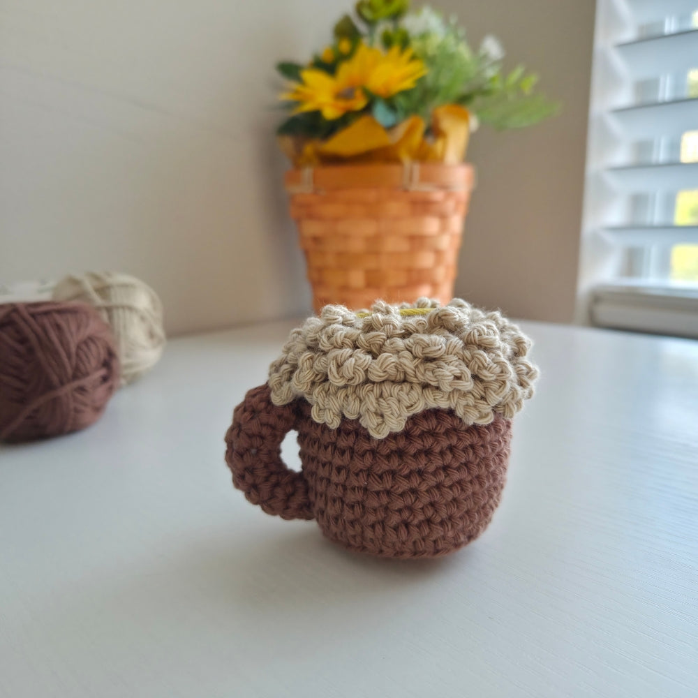 A crocheted mug-shaped item in medium brown with a thick, textured cream-colored top, displayed on a white tabletop with a blurred background showing a basket of yellow flowers and yarn balls.