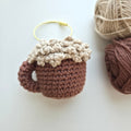 A crocheted mug-shaped ornament or charm in warm brown and cream, with a small yellow loop at the top, photographed on a light neutral background with balls of yarn nearby.