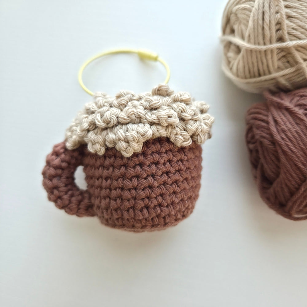 A crocheted mug-shaped ornament or charm in warm brown and cream, with a small yellow loop at the top, photographed on a light neutral background with balls of yarn nearby.