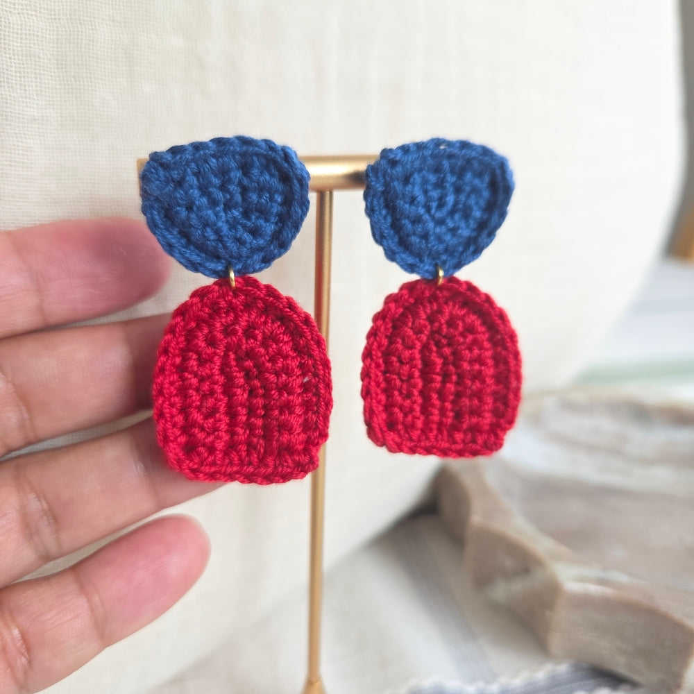 Crocheted earrings with blue and red shapes held by a hand against a neutral background