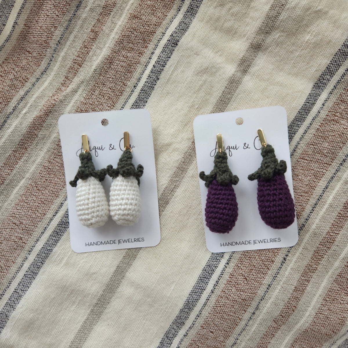 Pair of white and purple handmade crochet earrings on cards with a striped fabric background