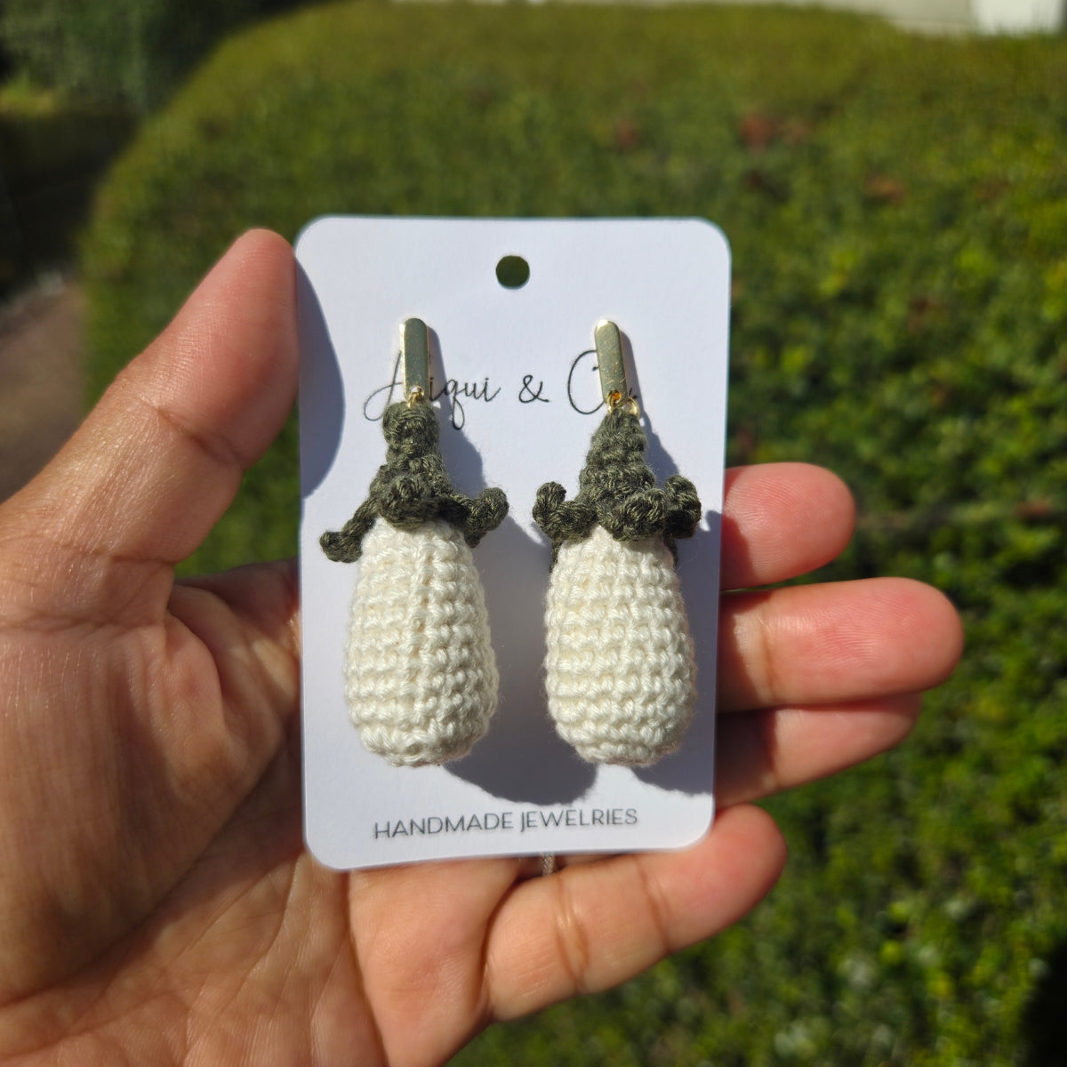 White eggplant earrings on a card held by a hand with a green outdoor background