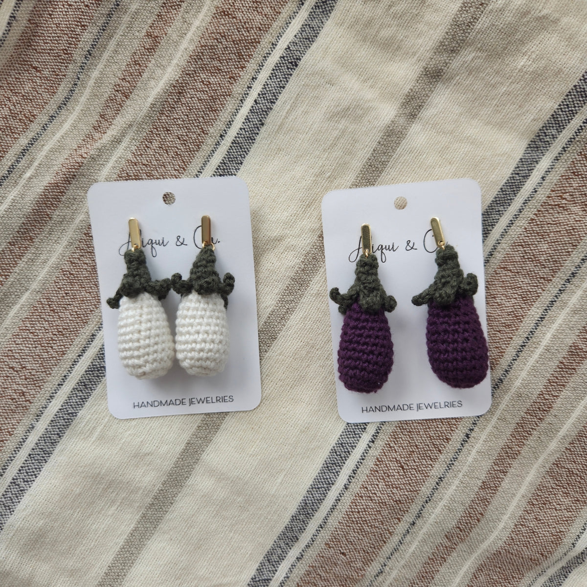 Two pairs of handmade eggplant earrings on cards with a striped fabric background