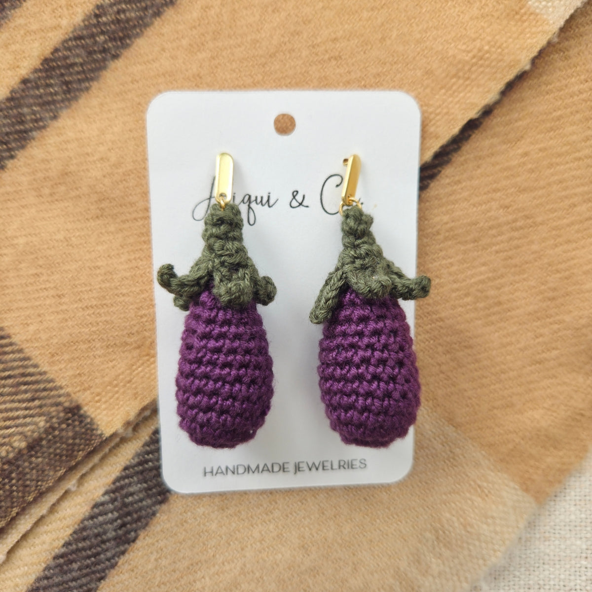 Crocheted eggplant earrings on a card with 'Aliqui & Co' branding, placed on a plaid fabric background.
