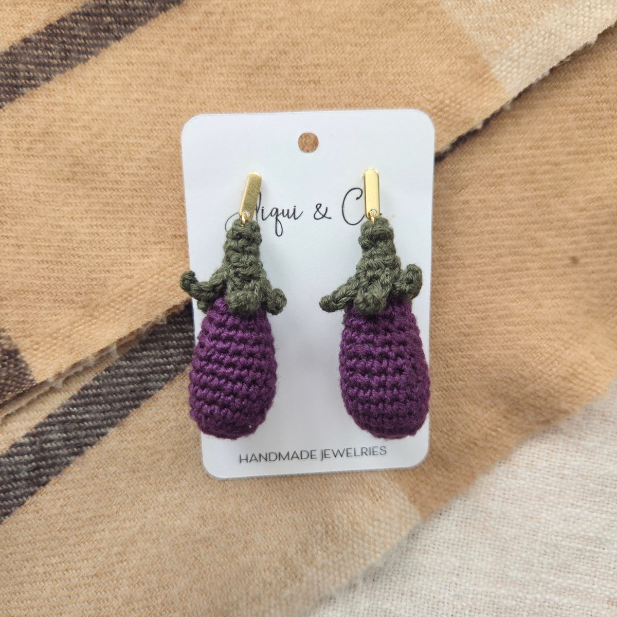 Crocheted purple eggplant earrings on a card with 'Aliqui & Co' branding, displayed on a textured fabric background.