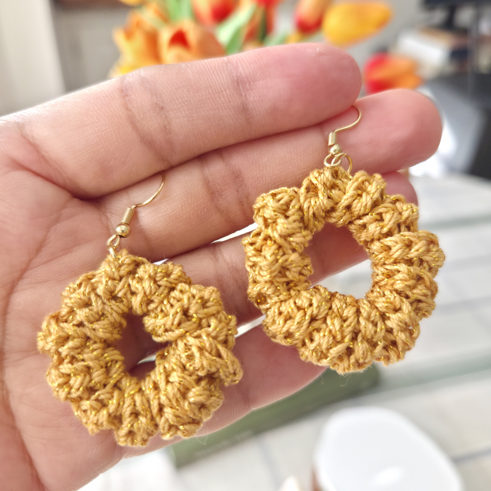 Hand holding a pair of gold crochet earrings with a blurred floral in background.