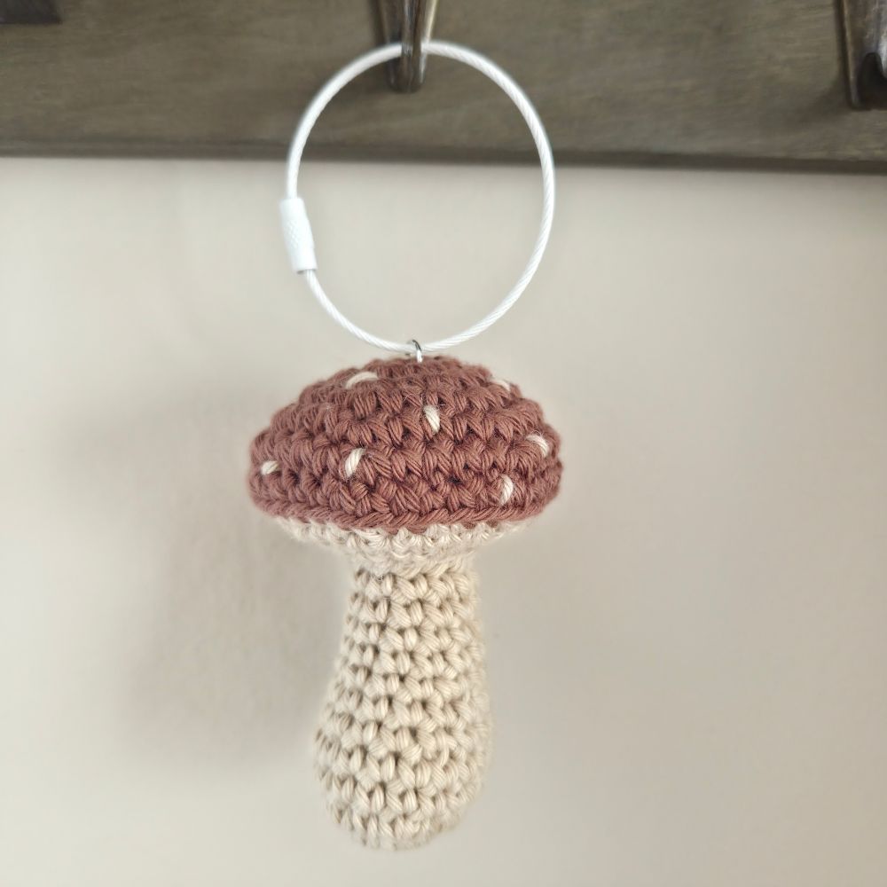 A crocheted mushroom-shaped hanging ornament or keychain with a clove brown cap and cream stem, attached to a white circular ring.