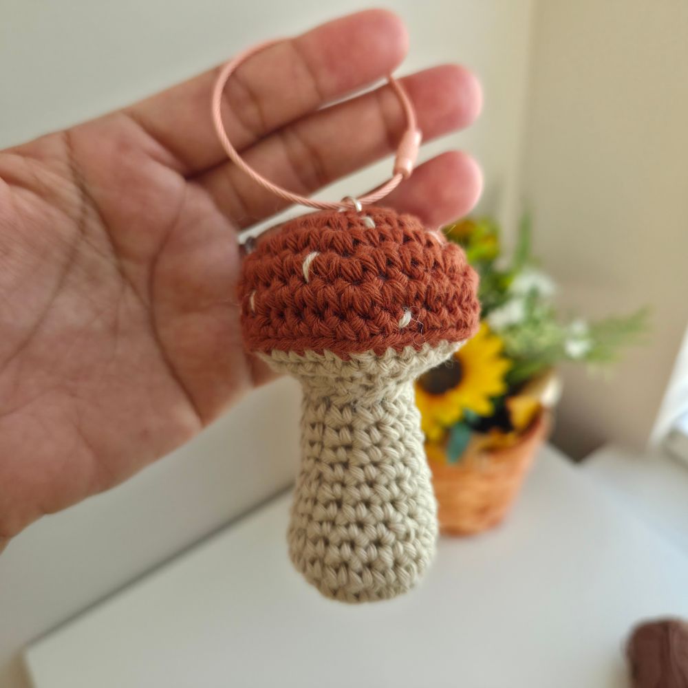 A hand holds a small crocheted mushroom charm with a terracotta brown cap, beige stem, and a light pink elastic loop, against a softly blurred indoor background.