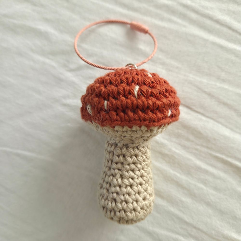 A handmade crocheted mushroom ornament with a terracotta orange cap, white spots, and a beige stem, hanging from a peach-colored elastic band on a white fabric background.