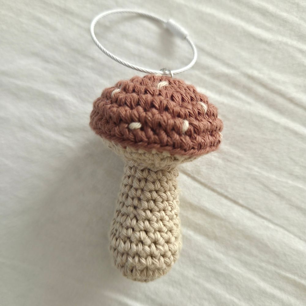 A crocheted mushroom ornament with a brown cap, beige stem, and white stitching details, hanging from a silver metal ring on a white fabric background.