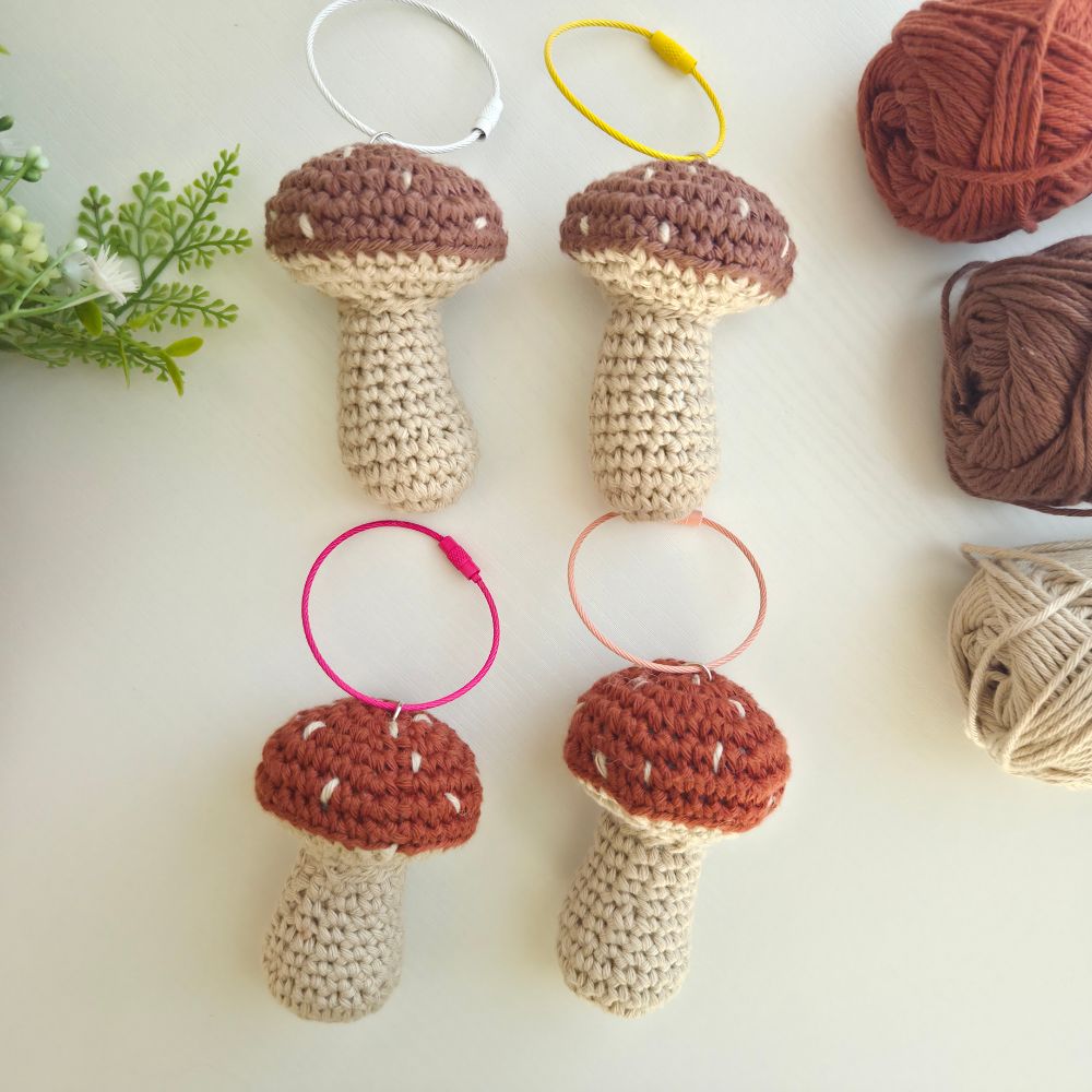 Four crocheted mushroom-shaped hair ties arranged on a light surface, each with a different colored key ring holder (white, yellow, pink, and peach). The image is styled with yarn balls and green leaves in the corners.