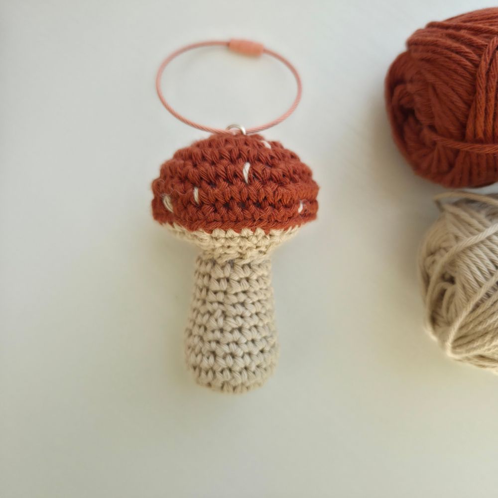 A crocheted mushroom-shaped bag charm or ornament with a rust-orange cap, cream stem, and a peach loop keychain holder at the top, shown on a light background with yarn balls partially visible at the edges.