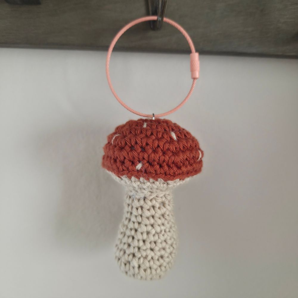 A crocheted mushroom-shaped ornament or keychain with a terracotta brown cap, cream stem, and a light pink ring, hanging against a plain light gray wall.