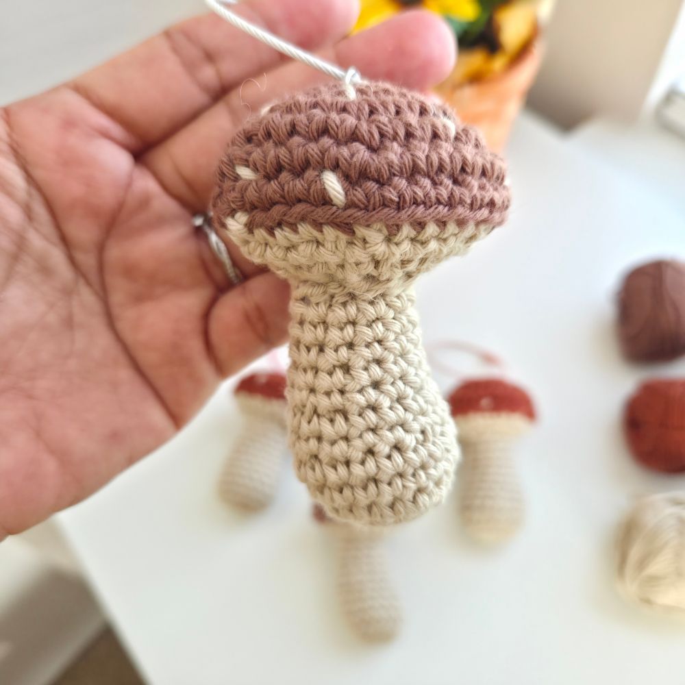 A handmade crocheted mushroom ornament held in a person's hand. The ornament features a textured brown cap with white stitch details and a cream-colored stem.