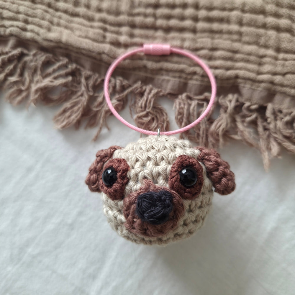 A handmade crocheted pug dog keychain or bag charm with a pink wire ring, resting on a white fabric surface with a brown fringed textile in the background.