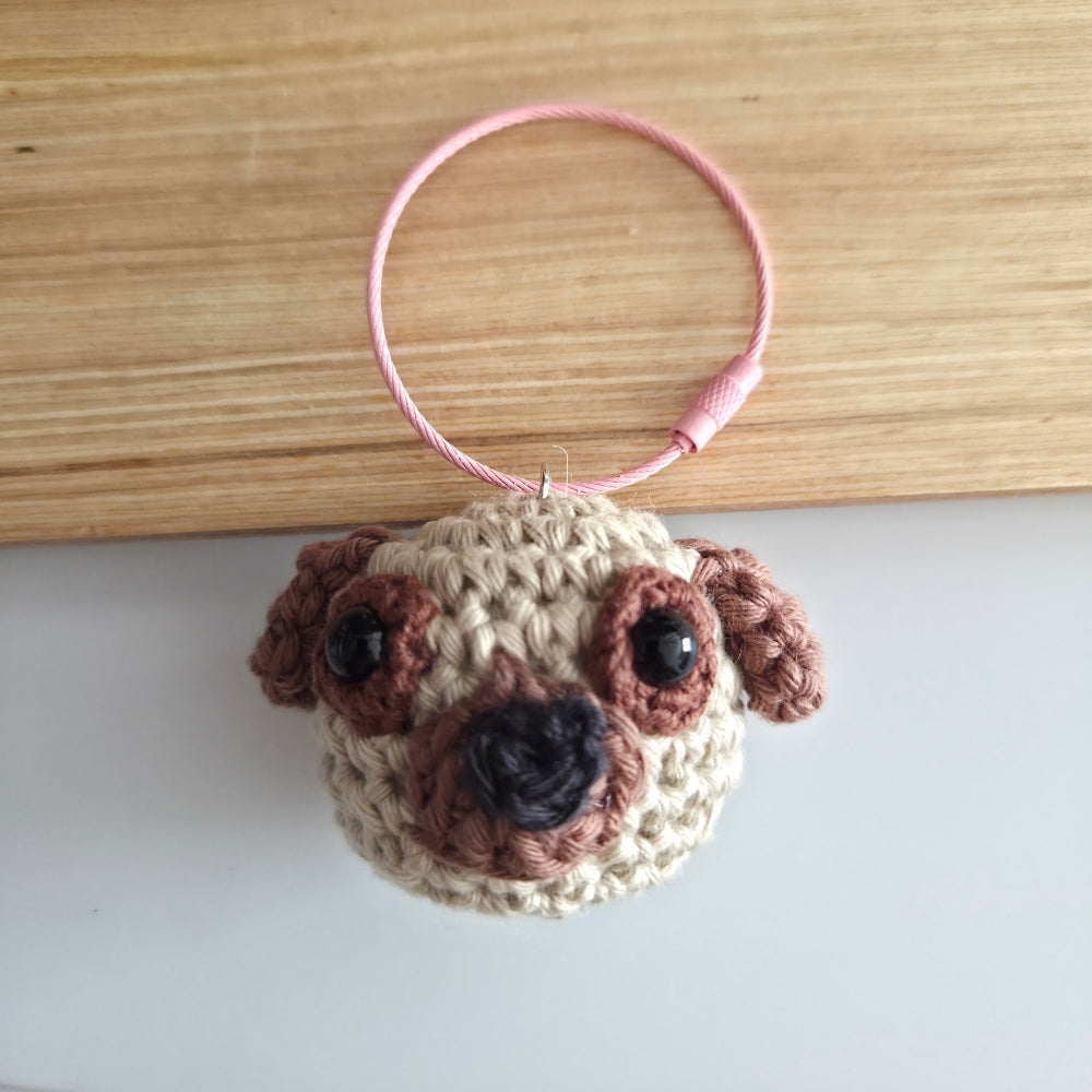 A crocheted pug face keychain or bag charm with a pink keyring loop, resting on a light wood surface against a white background.