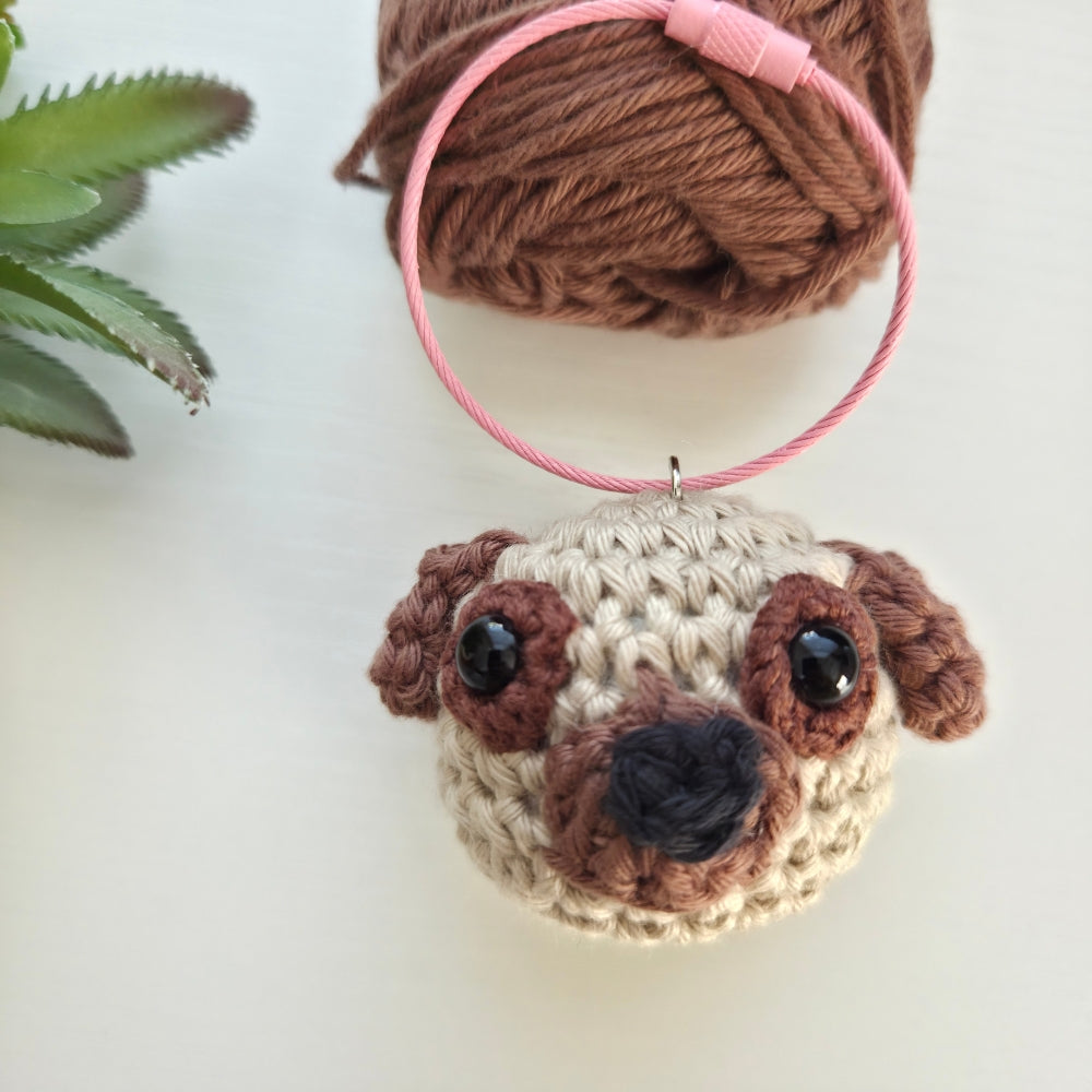 A crocheted pug dog head ornament or keychain charm with a pink hanging loop, shown on a light background with a ball of brown yarn and a small succulent in the background.