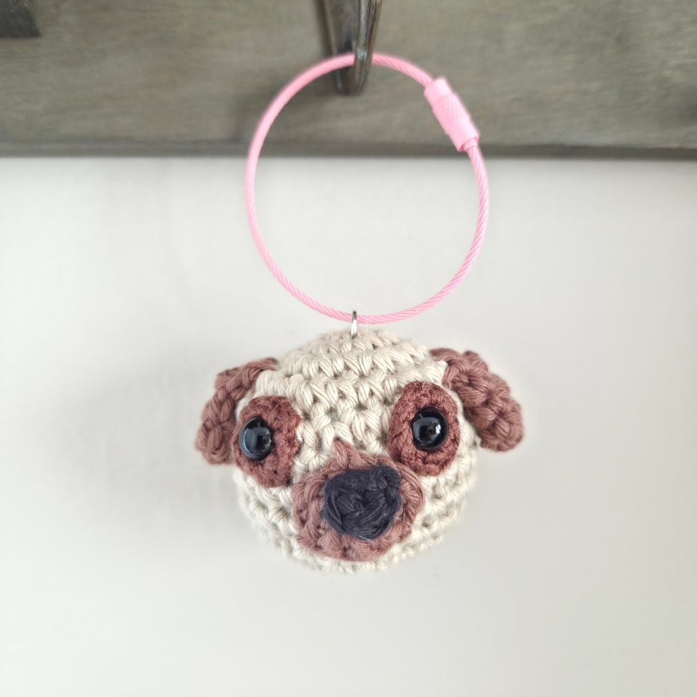 A crocheted pug dog head keychain or bag charm with a pink wire loop, hanging against a white background.