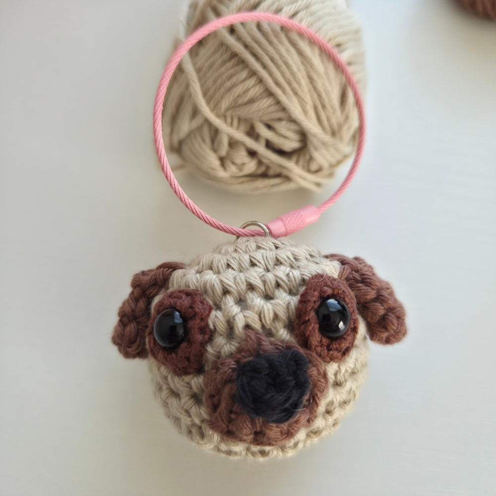 A crocheted pug dog keychain charm with a pink keyring, shown on a light off-white background with a ball of beige yarn behind it.