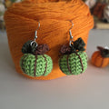 A pair of handmade crochet pumpkin earrings with light green bodies, dark green leaves, and brown stems, hanging in front of a large orange yarn ball.