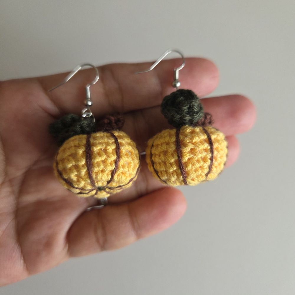 A pair of handmade crochet pumpkin earrings held in a person's hand. The earrings feature yellow and brown crocheted pumpkins with dark green tops and silver fishhook wires.