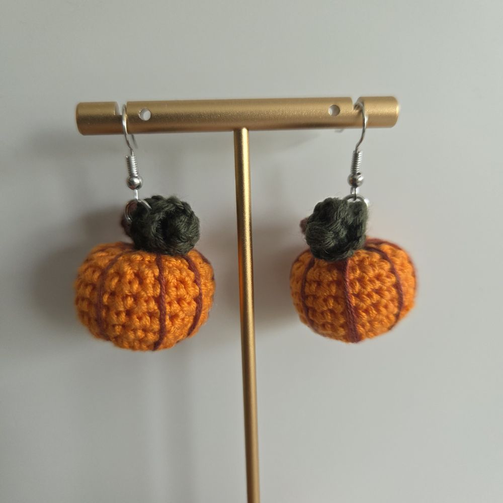 A pair of handmade orange crocheted pumpkin earrings with dark green stems, hanging from silver fishhook wires on a gold-colored T-bar display stand against a plain light gray background.