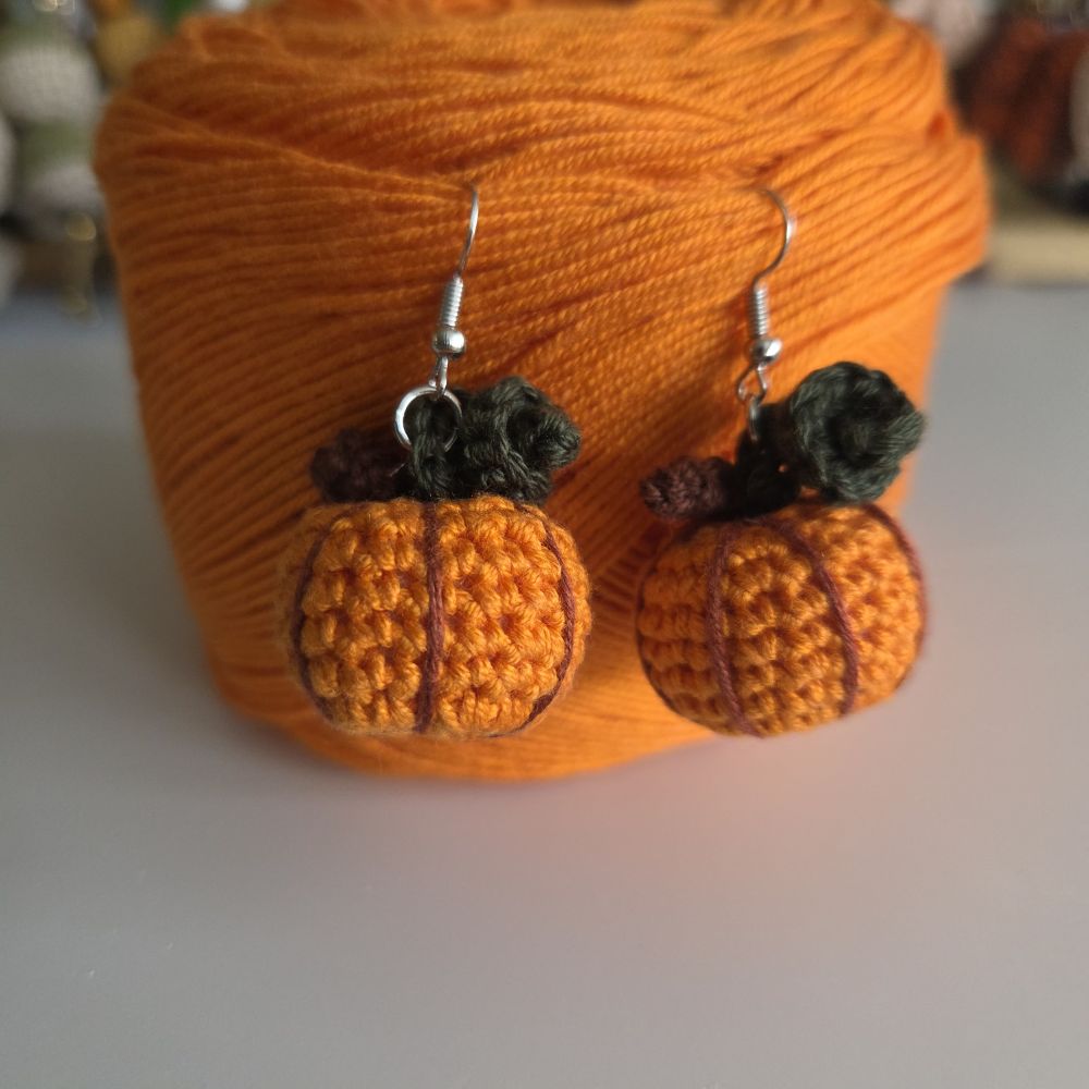 A pair of handmade crochet pumpkin earrings with dark brown stems and silver fishhook wires, displayed in front of a large spool of orange yarn.
