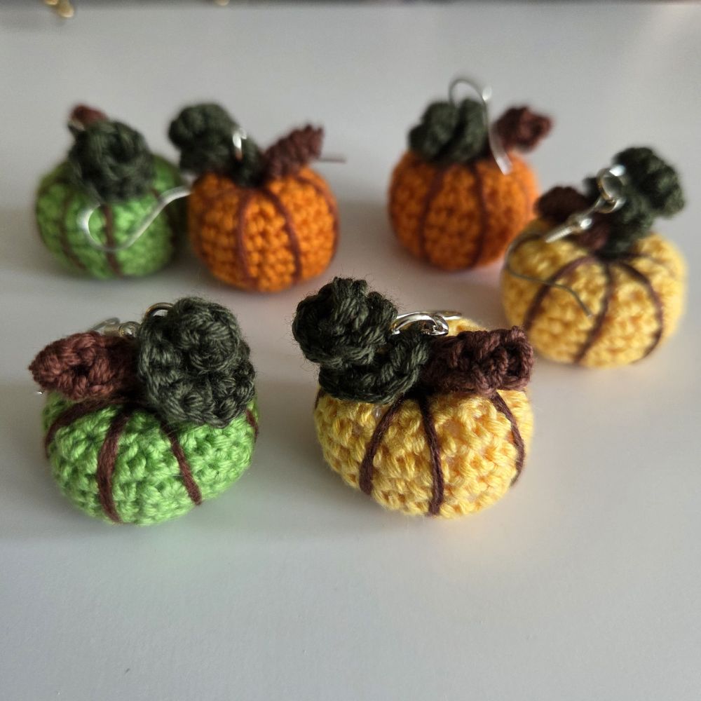 Six small crocheted pumpkin charms in green, orange, yellow, and brown, each with a dark green stem and silver metal hooks, arranged on a plain light background.