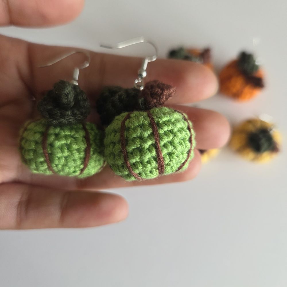 A pair of handmade green crochet pumpkin earrings held in a person's hand, with additional colorful crochet pumpkins blurred in the background against a plain light surface.