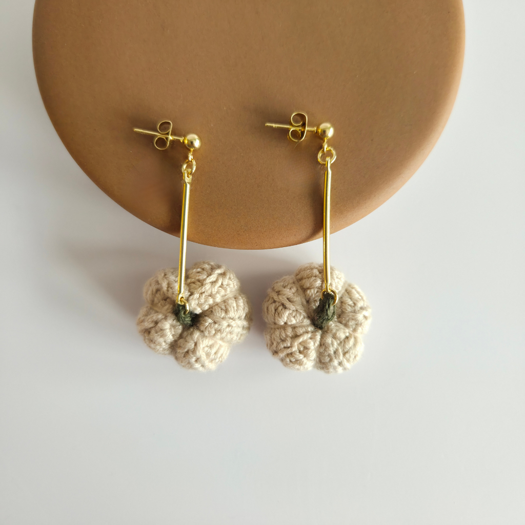 Pair of light tan crochet earrings with gold bar on a beige stand.