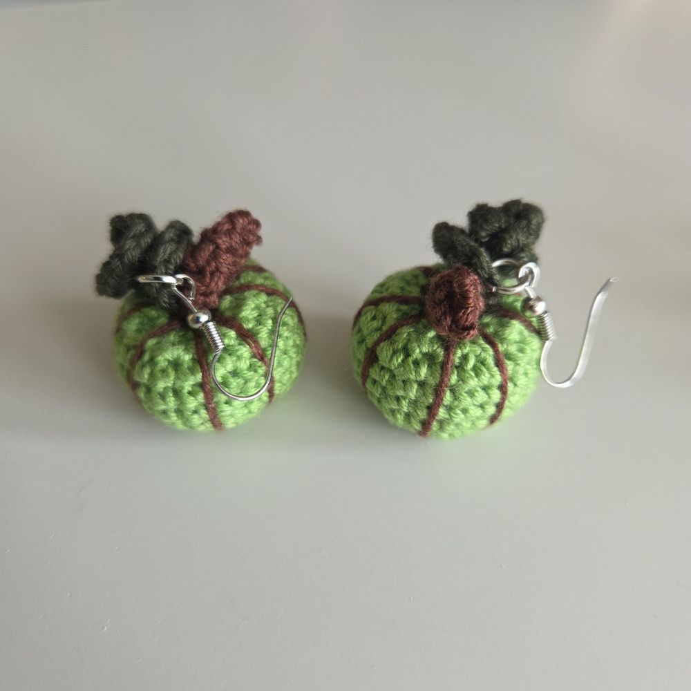 A pair of handmade crochet pumpkin earrings with green yarn bodies, brown stitched details, dark green stems, and silver fishhook ear wires, displayed on a neutral light gray background.