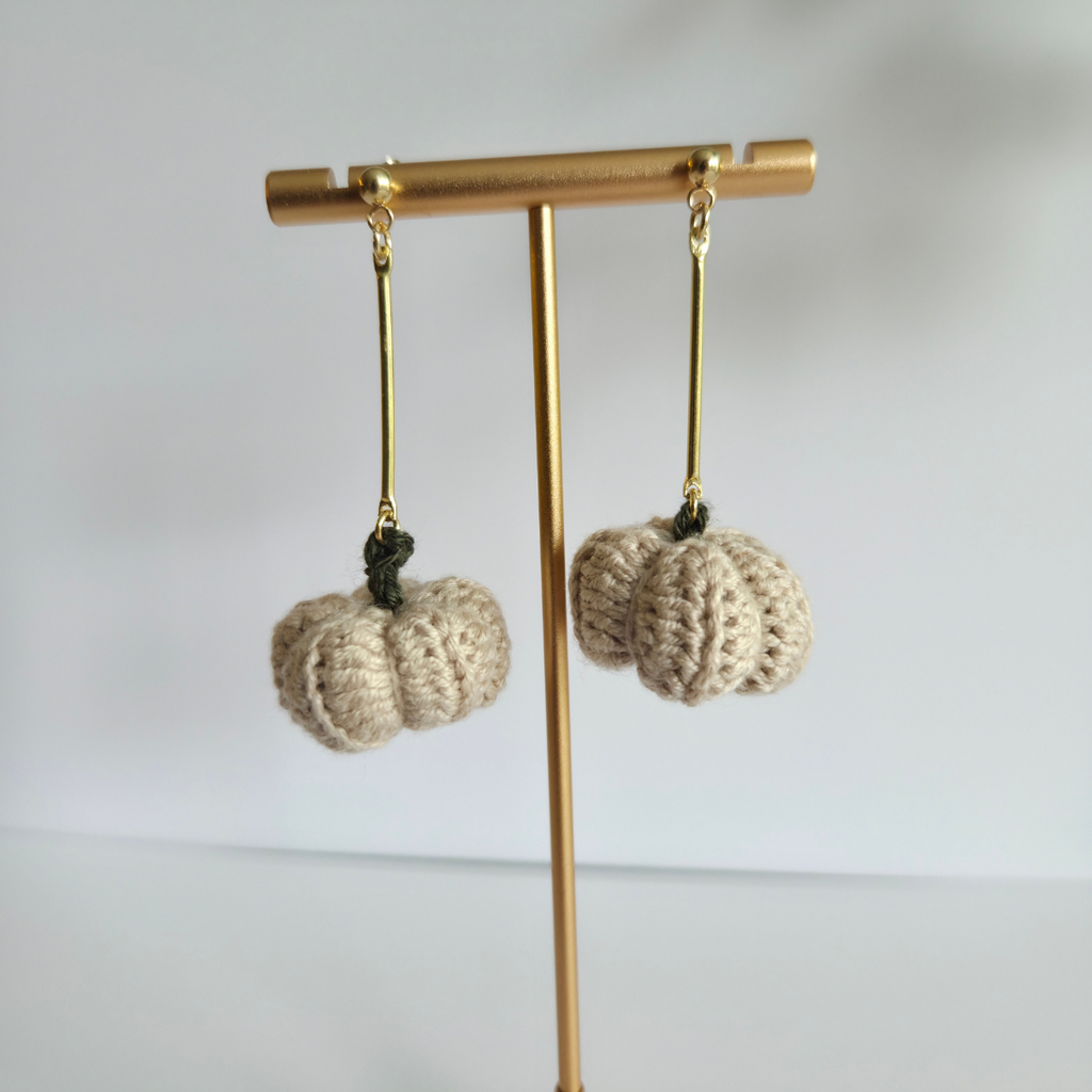 Crocheted beige pumpkin earrings on a gold earring stand against a white background