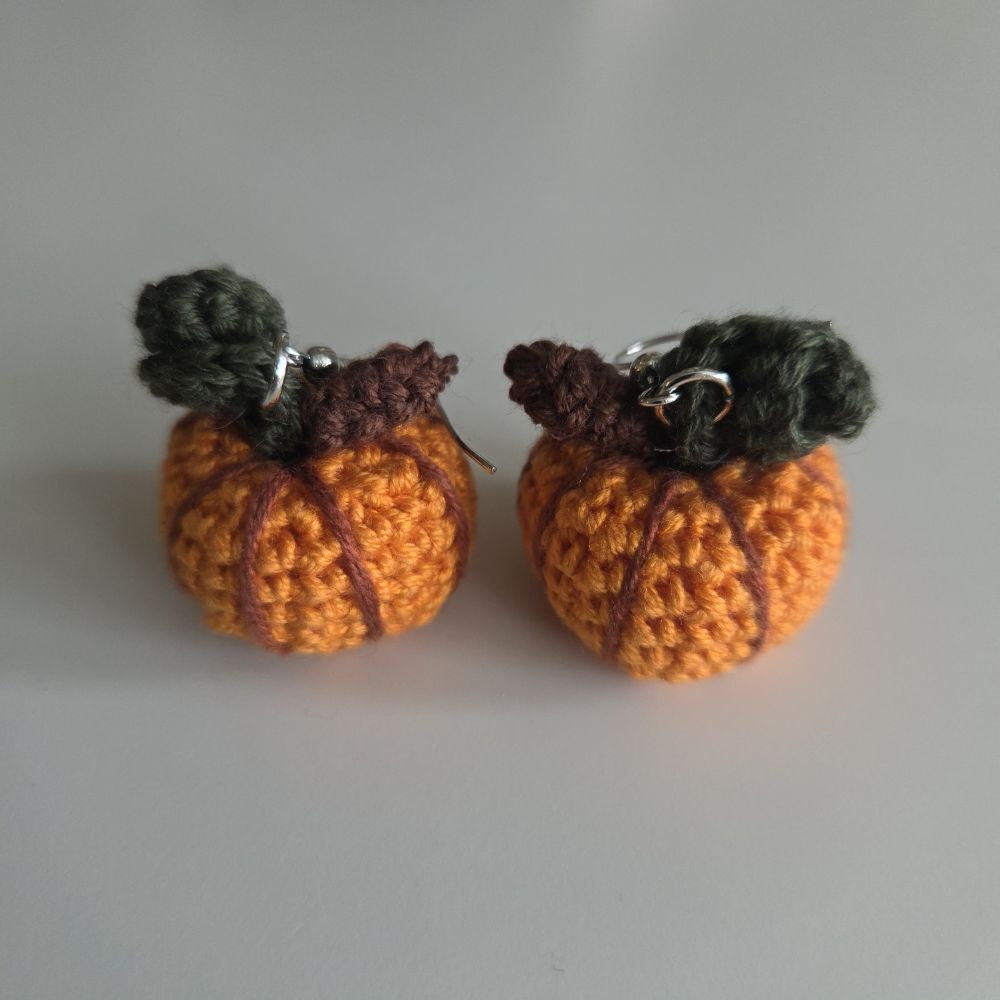 A pair of handmade crochet pumpkin earrings with orange bodies, dark brown stems, and dark gray leafy tops, shown on a neutral gray background.