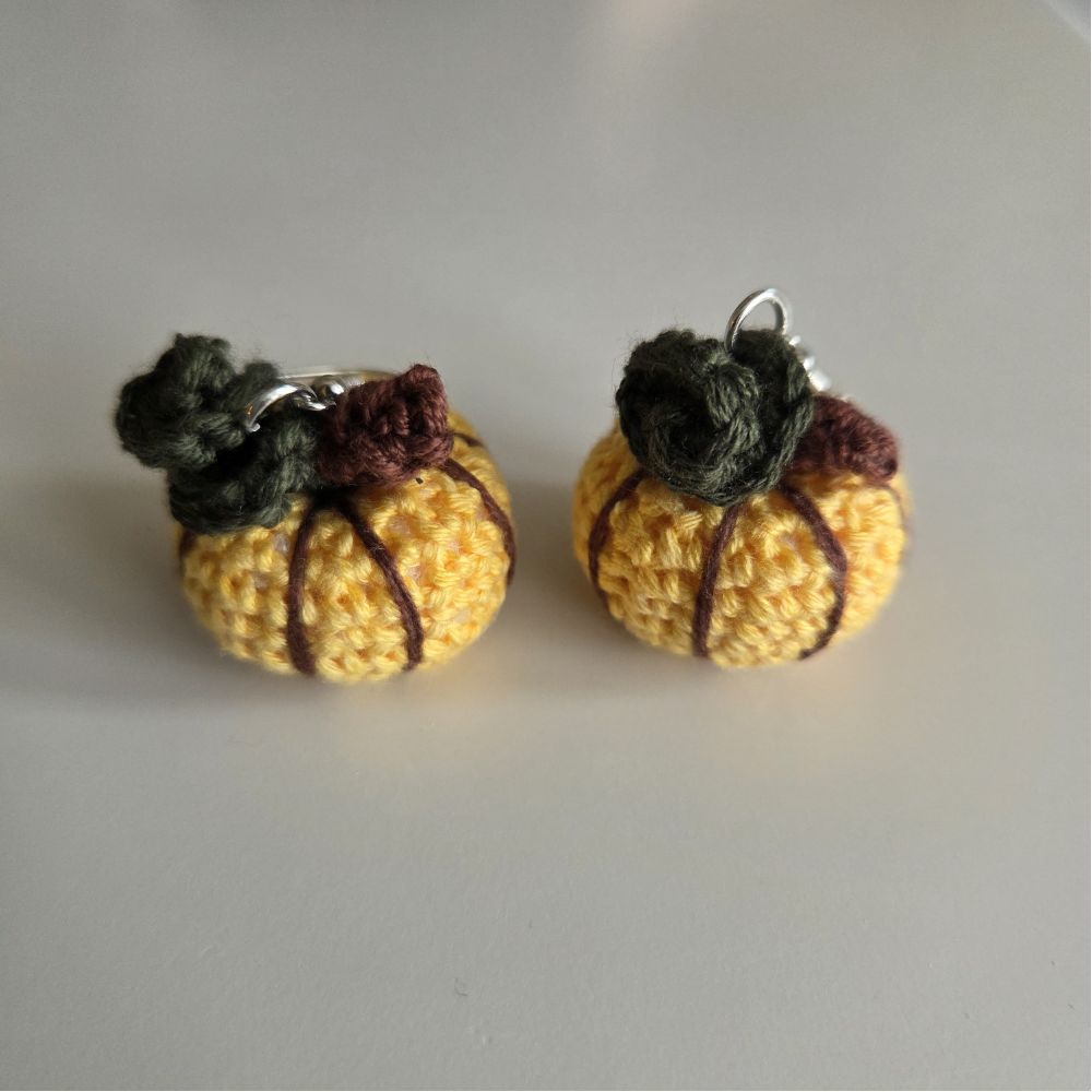 Two small crocheted pumpkin-shaped earrings with silver hooks, featuring yellow bodies, dark green stems, and dark brown accents, displayed on a neutral gray background.